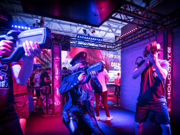 A Women In Uniform Pointing a Gun along with two men with guns and electronic gadgets over their eyes in a purple lit room