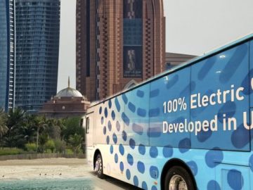 A Bus With Text That Reads 100 percent Electric Developed in UAE with two towers and a dome in the background