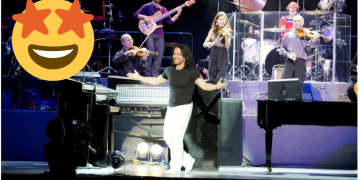 A Man Standing on a performace stage with his open arms with a muscial orchestra arranagement in the background