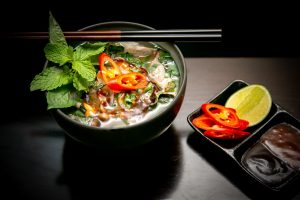Soup in a black bowl pair of chopsticks with garnishing of mint, chilly slices and other ingredients
