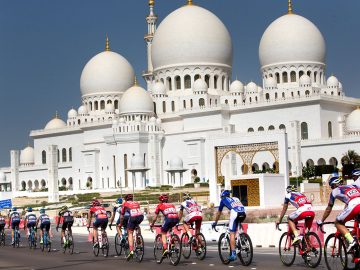 Cyclists riding in colorful jerseys with a white marble mosque in the background