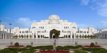 A wgite marble palace like structure under a blue sky in the background with green garden in the foreground