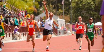 Athletes running on a red track with a winner in white vest rasing his one hand up in motion