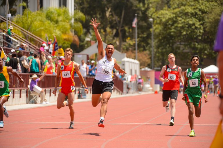 Athletes running on a red track with a winner in white vest rasing his one hand up in motion