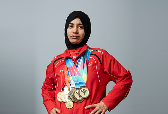 An athelete in red jacket and black head scarf posing with meadls around her neck